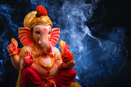 A vibrant Ganesh idol adorned with marigold flowers, set against a dark background with swirling blue smoke.